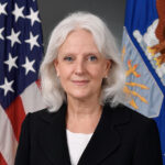 Woman wearing black suit against backdrop of American flags.